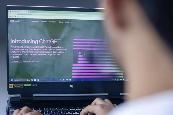 Eine Person sitzt vor einem Laptop.  Auf dem Laptop-Bildschirm befindet sich die Startseite des Chatbots ChatGPT für künstliche Intelligenz von OpenAI.