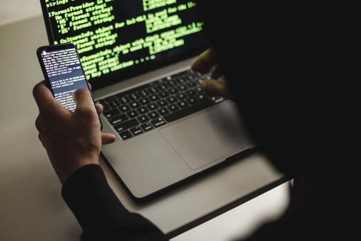 Ein Hacker tippt auf einem Apple MacBook-Laptop, während er ein Telefon in der Hand hält.  Beide Geräte zeigen Code auf ihren Bildschirmen an.