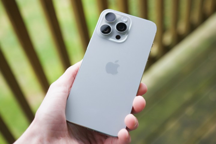 Jemand hält draußen auf einer Terrasse ein iPhone 15 Pro Max und zeigt die Rückseite in der Farbe „Naturtitanium“.